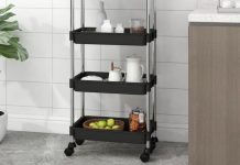 Black three-tier trolley in a modern kitchen, holding various items such as jars and fruit, next to a potted plant and a kitchen cabinet.