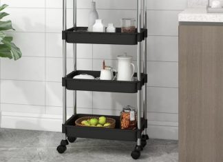 Black three-tier trolley in a modern kitchen, holding various items such as jars and fruit, next to a potted plant and a kitchen cabinet.