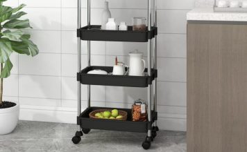 Black three-tier trolley in a modern kitchen, holding various items such as jars and fruit, next to a potted plant and a kitchen cabinet.