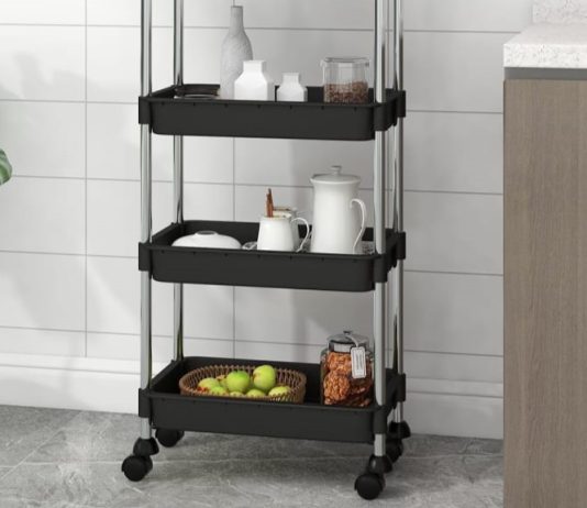 Black three-tier trolley in a modern kitchen, holding various items such as jars and fruit, next to a potted plant and a kitchen cabinet.